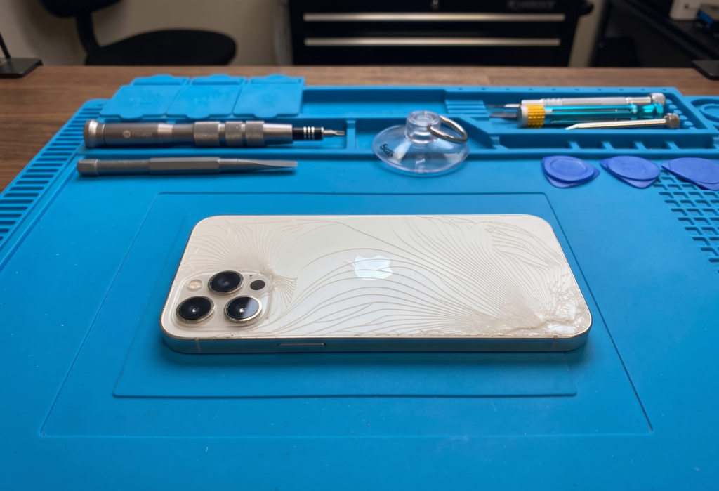 A white iPhone with a severely shattered back glass panel lying on a professional blue silicone repair mat at a mobile technician workstation.
