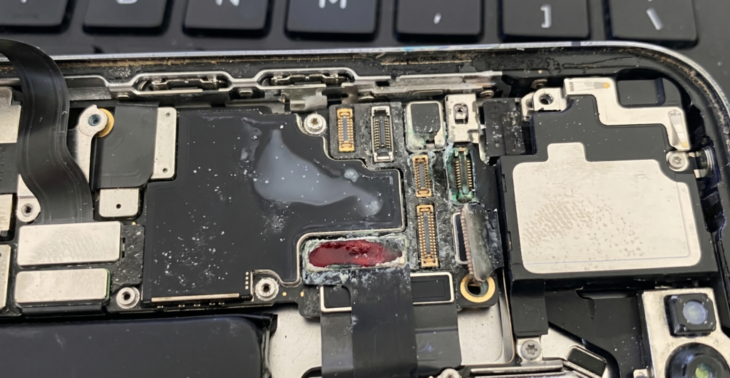 A high-resolution landscape close-up of an opened iPhone internal circuit board showing severe water damage, white mineral deposits, and green oxidation on the connectors and motherboard components (Water Damage Inside iPhone).